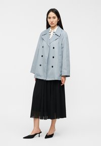 Ανοιχτό μπλε διπλόφρακτο παλτό με μαύρα κουμπιά, συνδυασμένο με μαύρη πλισέ φούστα και μαύρα μυτερά mules. Λείο υφή, κλασικός σχεδιασμός.