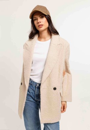 Woman wearing beige textured coat, white shirt, blue jeans, and brown cap against a plain light background.