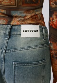 Jeans de mezclilla azul deslavado con un parche de cuero blanco con la etiqueta "URTRN" en la parte trasera. Presenta un diseño clásico de cinco bolsillos y costuras sutiles.