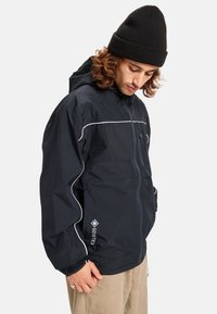 Zwarte Gore-Tex jas met witte piping, trekkoordcapuchon en elastische manchetten. Voorzien van een ritssluiting en logo op de onderrand.
