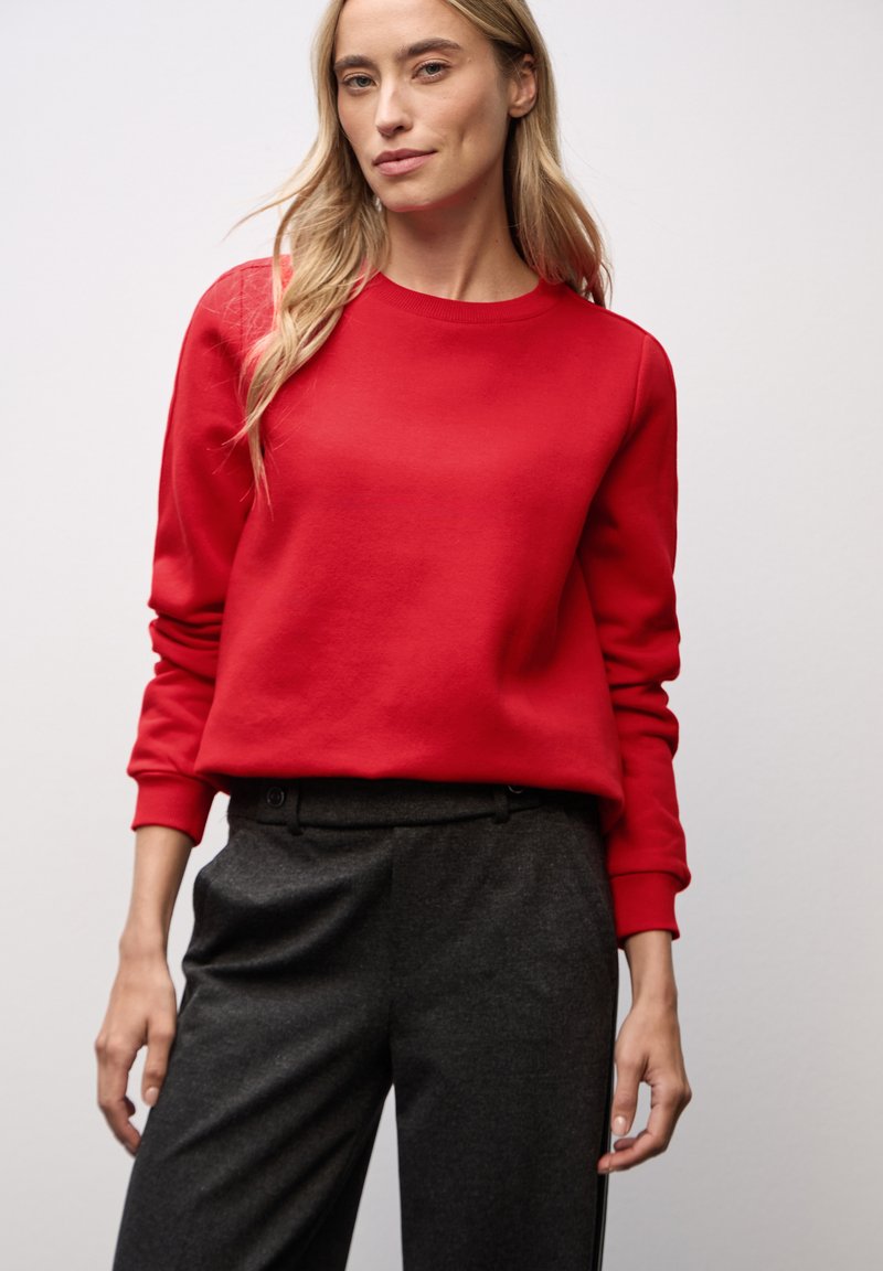 Rotes Sweatshirt mit Rundhalsausschnitt und langen Ärmeln, kombiniert mit dunkelgrauen, maßgeschneiderten Hosen, die eine glatte Textur und einen entspannten Fit zeigen.