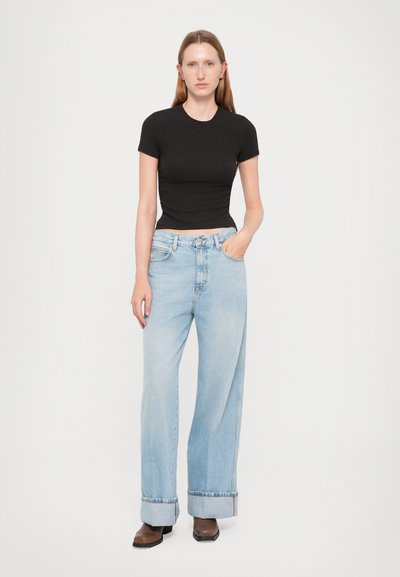 Young woman stands wearing black fitted short-sleeve top, high-waisted wide-leg light blue jeans with cuffs, and brown boots on white background.