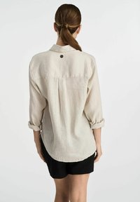 Beżowa lniana koszula z podwiniętymi rękawami, zaokrąglonym dołem i małym logo z tyłu. Teksturowany materiał nadaje lekki wygląd.