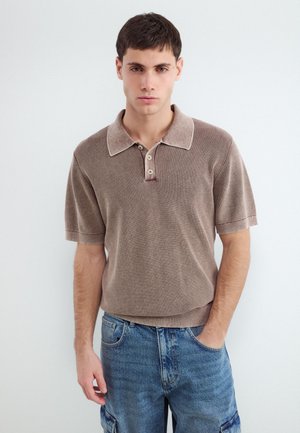 Junger Mann mit kurzen dunklen Haaren, der ein beiges kurzärmeliges Strick-Poloshirt und blaue Denim-Cargoshorts trägt und mit einer Hand in der Tasche steht.