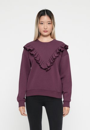 Lilla genser med V-formet volangdesign langs halsen, lange ermer og innsvinget midje. Laget av myk stoff.