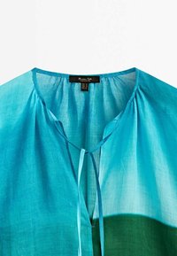 Blouse bleu clair avec encolure ronde et détail de nœud à l'avant, montrée de la partie supérieure de la poitrine jusqu'au col.
