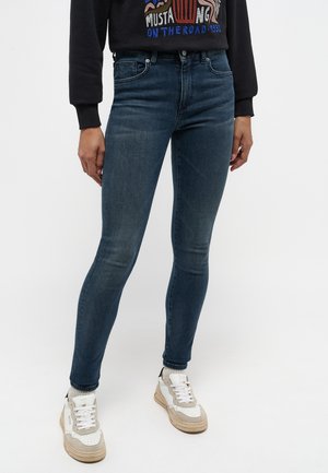 Jeans en denim bleu slim avec un léger délavage, cinq poches, matériel métallique et une texture lisse. Associés à des baskets blanches.