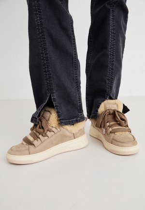 Beżowe sneakersy z grubymi brązowymi sznurówkami i puszystą wyściółką, noszone z czarnymi spodniami z rozciętymi nogawkami powyżej kostki.