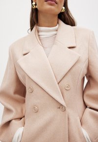 Svetloružový sako na dvojité zapínanie z mäkkej tkaniny, s veľkými výstrihmi a dekoračnými gombíkmi, s bielymi pletenými akcentmi na rukávoch.