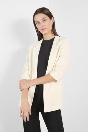 Cremefarvet blazer med en afslappet pasform, enkelt knaplukning, opsmøgede ærmer og glossy stof. Bæres over en tætsiddende sort bluse.