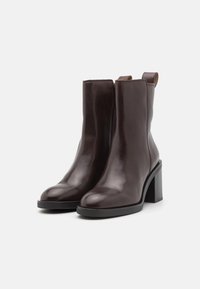 Botas de tobillo de cuero marrón con textura suave, puntera redonda, cremallera lateral y tacón alto grueso negro. Diseño minimalista sin adornos.