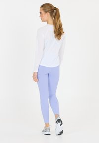 Långärmad vit atletisk topp med rund halsringning, i kombination med ljuslila leggings. Modellen bär grå sneakers med svart och vita detaljer.