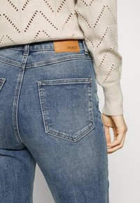 Jeans de mezclilla azul claro con un ligero desgastado, que presentan una etiqueta de cuero marrón en la cinturilla y dos bolsillos traseros. Suéter de punto texturizado arriba.