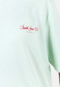 Camiseta de algodón color verde menta con mangas cortas, que presenta texto bordado en rojo "Sixth June Co. PARIS" en el lado izquierdo del pecho.