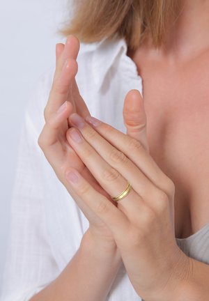 Gold ring with two smooth, parallel bands worn on a hand with natural nails, next to another hand in a relaxed position. Light background.