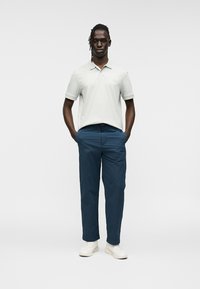 Ανοιχτό γκρι πόλο μπλουζάκι με κοντά μανίκια και γιακά· σκούρο μπλε παντελόνι· λευκά αθλητικά παπούτσια· το μοντέλο στέκεται με τα χέρια στις τσέπες.