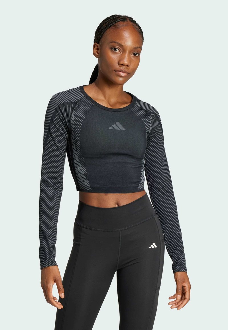 Crop top nero a maniche lunghe con texture orizzontale, impreziosito dal logo Adidas. Abbinato a leggings neri a vita alta, in tessuto liscio.