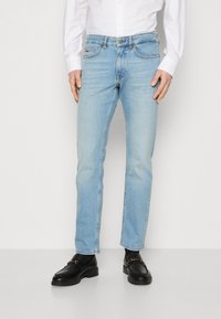 Jean en denim bleu clair avec une coupe droite, cinq poches et des surpiqûres contrastantes. Porté avec des mocassins en cuir noir et une chemise blanche.