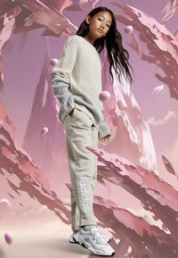 Světle béžový pletený svetr, béžové tepláky s logem "adidas" a bílé tenisky se šedými akcenty. Pozadí obsahuje abstraktní tvary.