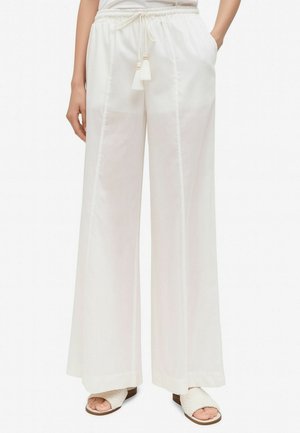 Donna che indossa pantaloni bianchi a gamba larga con cordoncini decorativi e sandali bianchi intrecciati con punta aperta, in piedi con le mani nelle tasche.