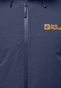 Veste bleu marine en tissu lisse et léger, dotée d'une fermeture éclair sur le devant, d'une capuche ajustable et d'un logo orange visible.