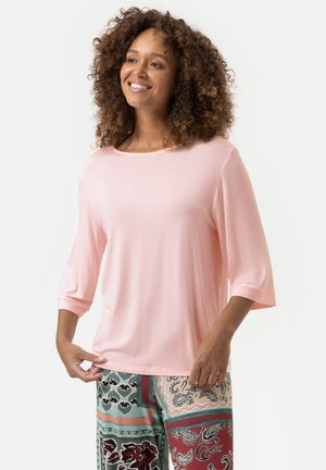Haut rose clair, ample, avec des manches trois-quarts et un col arrondi. Le tissu semble doux et lisse. Pantalons à motif visibles en dessous.
