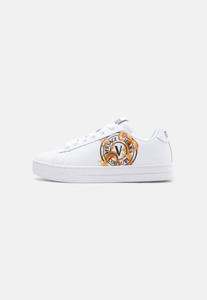 Biały niski sneaker z logo Versace Jeans Couture w kolorze złotym i czarnym po bokach oraz białymi sznurowadłami, pokazany na jednolitym tle.