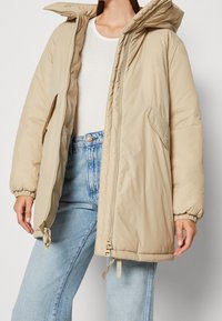 Chaqueta puffer beige con capucha, que cuenta con cierre de cremallera, puños elásticos y dos bolsillos laterales. La tela tiene una textura suave y aislante.