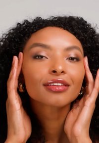 Femme aux cheveux bouclés touchant doucement son visage, portant des boucles d'oreilles en or en forme de créole et un maquillage naturel, sur un fond clair uni.
