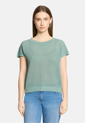 Jeune femme aux cheveux bruns raides portant un haut tricoté manches courtes vert clair et un jean bleu, debout devant un fond uni.