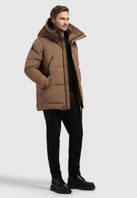 Hnedá pufková bunda s kapucňou, so zipsovým predným uzáverom, bočnými vreckami a textúrovanou podšívkou. V kombinácii s čiernymi nohavicami a masívnymi čiernymi čižmami.