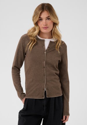 Femme aux longs cheveux blonds portant un cardigan marron boutonné sur un haut blanc et un pantalon noir, debout avec une main dans la poche.