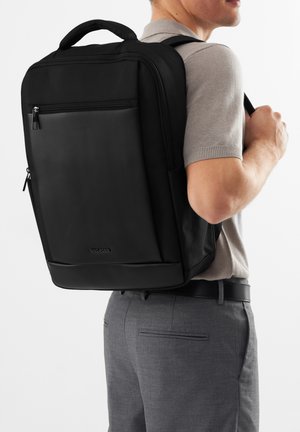 Uomo che indossa pantaloni grigi e una polo, che porta uno zaino nero con tasca frontale con cerniera e manico superiore.