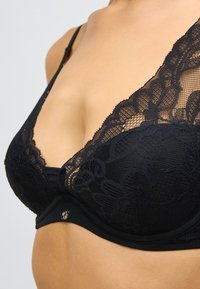 Melns mežģīņu krūšturis ar ziedu rakstiem, dziļu izgriezumu un gludu audumu. Lentes daļā ir dekoratīvs metāla elements.