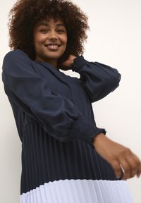 Tumši zilā pleitotā kleita ar baltu apakšmalu, ar garām pūkainām piedurknēm un savilkuma aprocēm, izgatavota no viegla auduma.