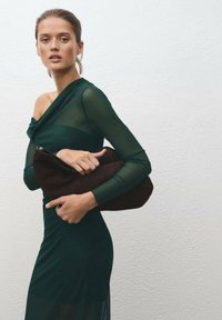 Geantă de mână de culoare maro închis, ținută în mâini, asociată cu o rochie ajustată de culoare verde închis, cu mâneci transparente și un design cu umerii la vedere.