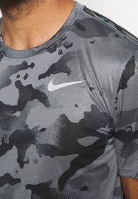Grå camouflage kortærmet atletisk skjorte lavet af glat stof, med en subtil glans og en lille hvid Nike-logo på venstre bryst.