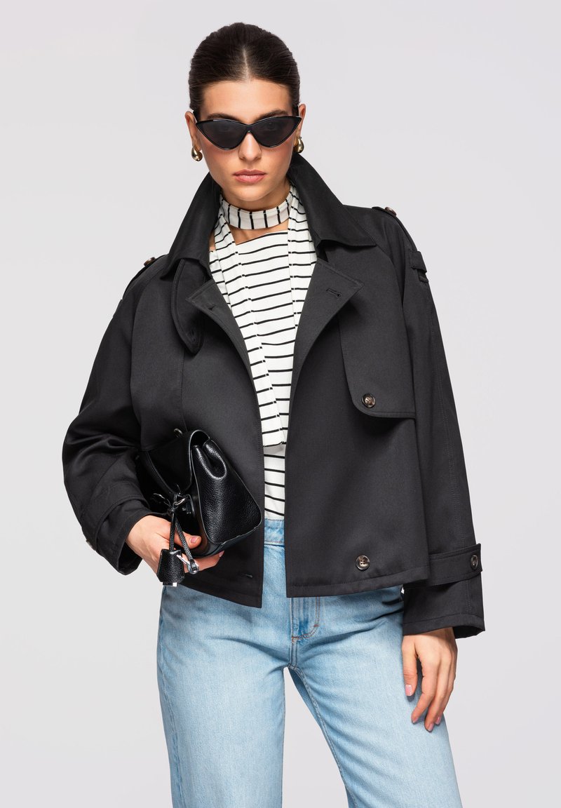 Femme portant une veste courte noire, un top rayé avec col montant, un jean bleu clair, des lunettes de soleil œil-de-chat, des boucles d'oreilles en or, tenant un sac à main noir.