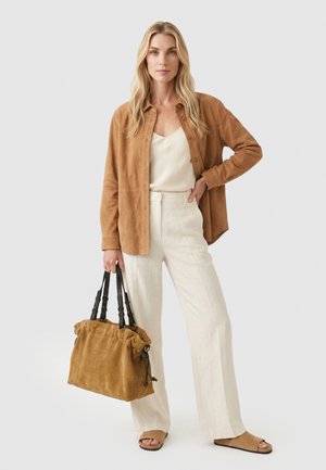 Blondine met een lichtbruine suède blouse, witte top, witte broek en lichtbruine sandalen, met een grote lichtbruine suède handtas met zwarte handvatten.