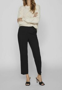 Kremowy dzianinowy sweter z teksturowanym wzorem w diamenty, zestawiony z czarnymi dopasowanymi spodniami i czarnymi sandałami na obcasie.