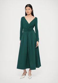 Haljina dugih rukava, dubokog V-izreza u zelenoj boji s uvijenim strukom i lepršavom suknjom. Glatki materijal, duljine do gležnjeva, u paru s bijelim cipelama na petu.