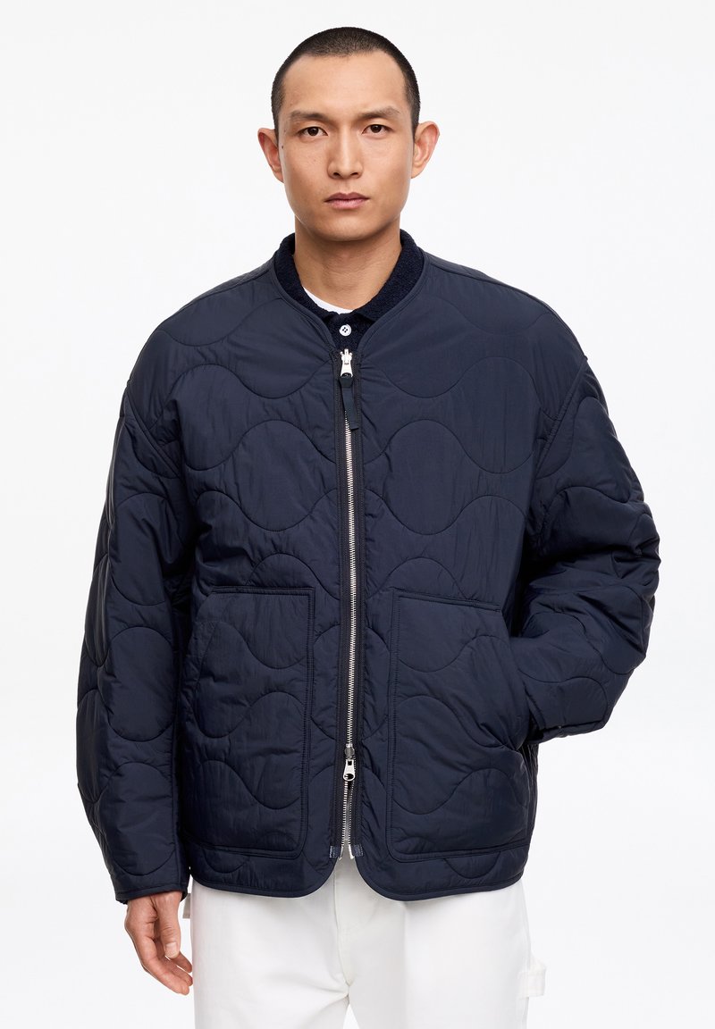 Hombre con chaqueta acolchada azul marino con bolsillos delanteros y cremallera plateada, combinado con pantalones blancos, de pie frente a un fondo liso.