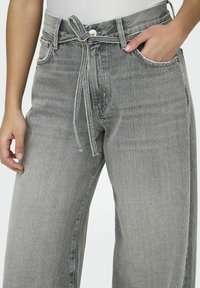 Grå denimbukser med høy midje, snøring og brede ben. Har frontlommer og en subtil falmet tekstur.