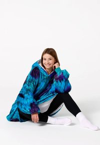 Modrá a fialová oversized kapucňová prikrývka s batikovaným vzorom, plyšová textúra, veľké rukávy, nosená cez čierne legíny a biele ponožky, sedacia póza.