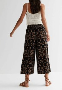Woman wearing black wide-leg pants with brown geometric patterns, white crochet tank top, and brown lace-up flat sandals, standing against white background.