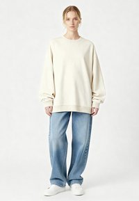 Krēmkrāsas oversized džemperis ar nolaižamām plecu līnijām, ribstaina apkakle un aproces, kombinēts ar gaiši zilām brīvi piegulošām džinsa biksēm un baltām sporta kurpēm.