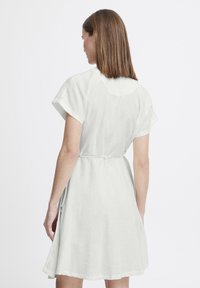 Lagani laneni haljina u boji off-white s kratkim rukavima, uvučenim strukom i raširenim rubom, koja prikazuje jednostavan i minimalistički dizajn.