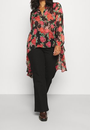 Person trägt eine schwarze, durchsichtige Bluse mit roten und pinken Blumen, schwarze Hose und braune Schuhe, geht vor einem weißen Hintergrund vorwärts.