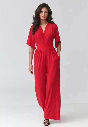 Kobieta z długimi ciemnymi włosami, ubrana w czerwony kombinezon z szerokimi nogawkami, krótkimi rękawami i czarne czółenka z odkrytymi palcami, stojąca na jednolitym tle.