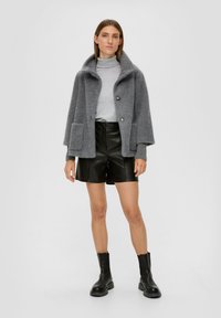 Veste oversized en herringbone gris avec un col haut, deux poches, associée à des shorts en cuir noir et des bottes chunky noires.
